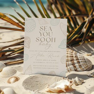 Może przedstawiać: Zaproszenie na baby shower z napisem "SEA YOU SOON baby" w stylowej czcionce. Zaproszenie jest ozdobione ilustracjami muszli i umieszczone na piaszczystej plaży z muszlami i liśćmi palmowymi.
