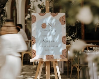 May include: A wooden easel displays a party sign with a light blue background and cookie illustrations. The sign reads "ONE Sweet Party" and welcomes guests to "Kai's First Birthday" on August 7, 20XX. The sign is surrounded by a blurred outdoor setting.