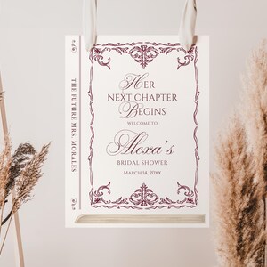 Puede incluir: Un letrero de despedida de soltera con el texto "Her Next Chapter Begins" y "Welcome to Alexa's Bridal Shower". El letrero tiene un borde decorativo burdeos y est&aacute; colgado de una cinta blanca. Tambi&eacute;n est&aacute; presente el texto "The Future Mrs. Morales".