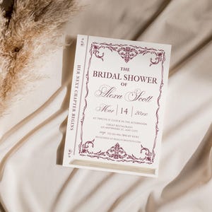 May include: A white invitation card with a burgundy floral border and text that reads "THE BRIDAL SHOWER of Alexa Scott Mar 14 202x". The card is designed to look like a book. The words "HER NEXT CHAPTER BEGINS" are printed vertically on the side.
