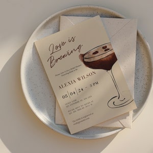 Puede incluir: Una invitaci&oacute;n beige para una despedida de soltera con el texto "Love is Brewing" y una ilustraci&oacute;n de acuarela de un c&oacute;ctel. La invitaci&oacute;n incluye detalles del evento, como la fecha, la hora y el lugar, y est&aacute; colocada sobre un plato moteado.