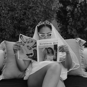 Könnte beinhalten: Schwarz-Weiß-Bild einer Person mit Schleier, Sonnenbrille und einem "Bride To Be"-Stirnband, die eine Zeitung mit dem Titel "Bachelorette Post" liest. Die Zeitung zeigt ein Foto einer Frau. Der Hintergrund umfasst Laub und Kissen.