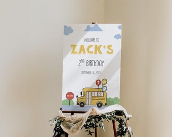 May include: A party welcome sign featuring the text "Welcome to Zack's 2nd Birthday" in yellow and black. The sign includes a yellow school bus illustration, balloons, clouds, and a stop sign. The sign is displayed on an easel.