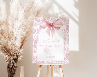 May include: A baby shower welcome sign in pink and white, displayed on an easel. The sign has a pink bow and floral pattern, with the text "Welcome To ALEXANDRA'S baby shower July 12, 2025."