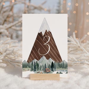 May include: A table number card featuring a watercolor illustration of a mountain scene. The number "3" is displayed in a cursive font. The mountain has a snow-capped peak and a wooden texture. The base of the card is supported by a wooden stand.