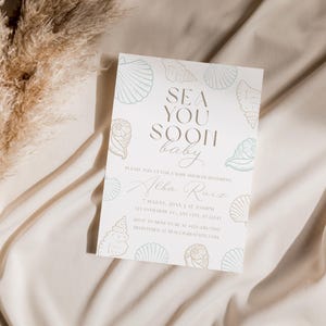 Może przedstawiać: Zaproszenie na baby shower z napisem "Sea You Soon Baby" w stylowej czcionce. Zaproszenie zawiera ilustracje muszli w miękkim odcieniu niebiesko-zielonym i zawiera szczeg&oacute;ły wydarzenia. Zaproszenie jest na białym tle.