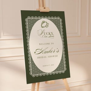 Op de afbeelding: Donkergroen bord op een houten ezel met de tekst "Lucky in Love" en "Welcome to Amber's Bridal Shower" in sierlijke letters. Het bord heeft een witte ovale kern met een kantachtige rand en een ringafbeelding.