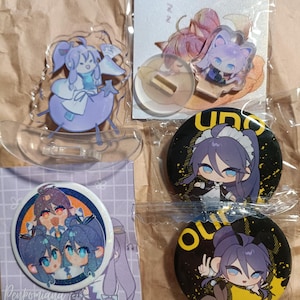 May include: A collection of anime-style merchandise including a clear acrylic standee of a character on a crescent moon, a small acrylic cat-ear character charm, and three circular pin-back buttons featuring illustrated characters with purple hair.