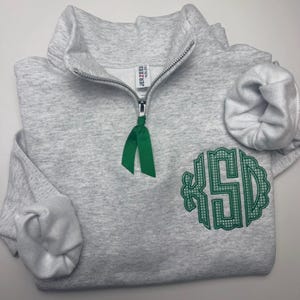 May include: A light gray heathered quarter-zip sweatshirt featuring a silver zipper pull adorned with a green ribbon. The garment displays a green embroidered monogram with the letters KSD in a scalloped circular frame on the lower right side.