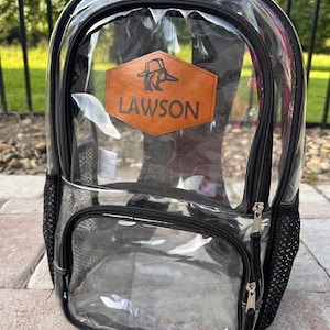 May include: Clear backpack with black trim and zippers. Features a front pocket, side mesh pocket, and a top handle. A brown leather patch with the name "LAWSON" and a duck logo is centered on the front. The backpack is designed for visibility and easy access.