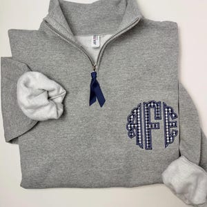 May include: A heather gray quarter-zip sweatshirt featuring a navy blue ribbon pull on the zipper. The front displays a large, circular monogram with the letters AFE in a navy blue and white gingham pattern, outlined in navy blue embroidery.