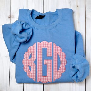 May include: A light blue sweatshirt with a pink gingham monogram. The monogram is in a scalloped circle. The sleeves are folded. The sweatshirt is folded on a white wooden surface.