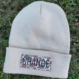 May include: A beige knit beanie featuring a rectangular patch on the cuff. The patch displays a camouflage pattern in shades of green, brown, and tan with the word CHANCE printed in bold black capital letters across the center.