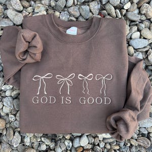 May include: A brown sweatshirt with the embroidered phrase "GOD IS GOOD" below four bow designs. The long-sleeved top is made of a soft material and is displayed on a bed of small rocks.