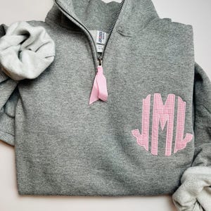 May include: Gray sweatshirt with a pink gingham monogram on the front. The sweatshirt has a zipper and a high neck.