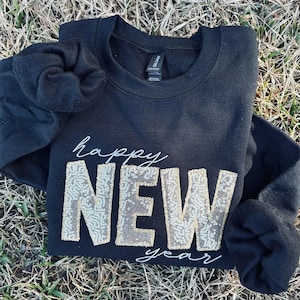 May include: Black sweatshirt with the words "happy NEW year" in white script and gold sequins. The sweatshirt is folded on a bed of dry grass.