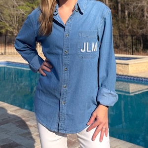 May include: A light blue denim shirt with long sleeves and a button-down front. The shirt has a chest pocket with the embroidered initials "JLM" in white. The sleeves are rolled up, and the shirt is paired with white pants.