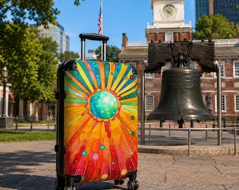 May include: A hard-shell rolling suitcase featuring a vibrant, multi-colored sunburst design with a central teal sphere, radiating orange, yellow, and red rays, accented by small circles, set against a background of a historic brick building and a bell.