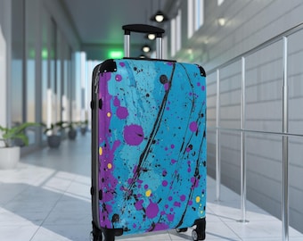 May include: A blue suitcase with a black handle and wheels. The suitcase has a colorful abstract design with splashes of purple, yellow, and black paint.