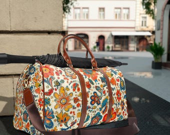 May include: A floral print duffel bag with brown leather handles and straps. The bag features a colorful pattern of flowers in orange, yellow, blue, and red on a cream background. A black umbrella rests on top of the bag. The bag has a shoulder strap.