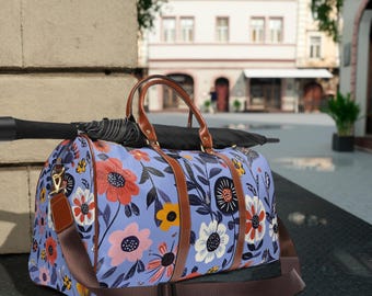 May include: A blue floral print duffel bag with brown leather accents and a long adjustable strap. The bag is sitting on a concrete surface.