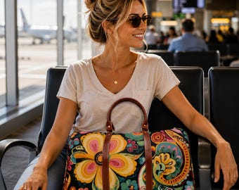 May include: A duffel bag featuring a vibrant, multi-colored floral and paisley pattern on a black background, accented with brown leather handles and trim. The bag has a structured rectangular shape with a top zipper closure and dual carry handles.