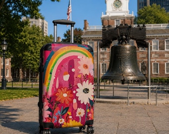 May include: A rolling suitcase featuring a vibrant pink background with a painted rainbow and various colorful flowers. The luggage has a retractable handle, four wheels, and black corner protectors, standing upright on a paved outdoor surface.