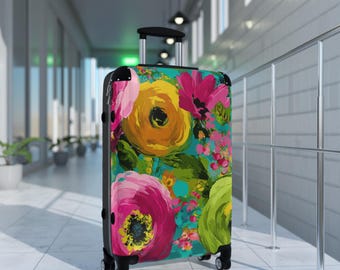 May include: A hard-shell rolling suitcase featuring a vibrant floral pattern with large yellow, hot pink, and lime green painted flowers on a teal background. The luggage has a black frame, black corner protectors, and a retractable silver handle.