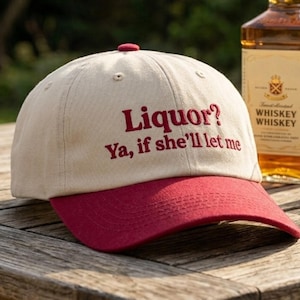 May include: A beige baseball cap with a red brim and the embroidered red text "Liquor? Ya, if she'll let me" on the front, positioned next to a glass bottle of whiskey and a clear glass containing a small amount of amber-colored liquid.