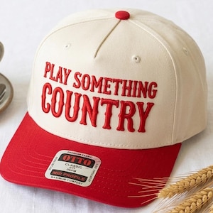 Op de afbeelding: Crème en rode baseballcap met de tekst "PLAY SOMETHING COUNTRY" in het rood geborduurd. Een kopje thee en tarwearen staan op de achtergrond, op een wit oppervlak.