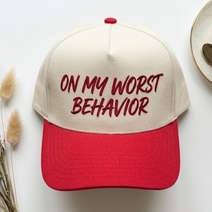 May include: A baseball cap with a cream-colored crown and a red brim. The phrase "ON MY WORST BEHAVIOR" is embroidered in red on the front. A small red button is on top of the cap.