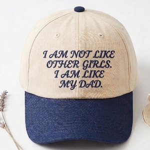 May include: A baseball cap with a beige crown and a navy blue brim. The cap features embroidered text that reads "I AM NOT LIKE OTHER GIRLS. I AM LIKE MY DAD."