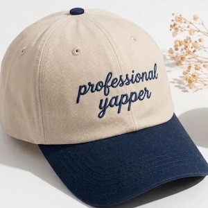 May include: A beige baseball cap with a navy blue brim. The cap features the embroidered text "professional yapper" in navy blue. The cap has a small navy blue button on top.