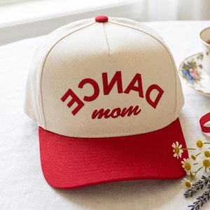 May include: A baseball cap with a cream-colored crown and a red brim. The cap features the words "DANCE mom" in red embroidered lettering. A teacup and flowers are in the background.