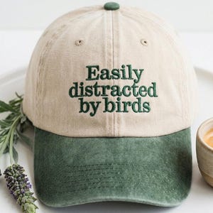 Może przedstawiać: Beżowa i zielona czapka z daszkiem z haftowanym napisem "Easily distracted by birds". Czapka ma zielony daszek i zielony guzik na górze. Mała filiżanka kawy stoi obok czapki.