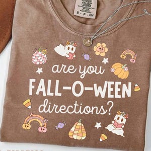 Könnte beinhalten: Braunes T-Shirt mit dem Text "are you FALL-O-WEEN directions?" und Halloween-Motiven. Das Design umfasst Geister, Kürbisse, Regenbogen und Süßigkeiten. Eine silberne Halskette liegt auf dem T-Shirt.