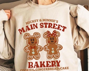 May include: A beige sweatshirt with a graphic of Mickey and Minnie Mouse dressed as gingerbread people. The text reads "Mickey & Minnie's Main Street Bakery Cookies Gingerbread Cake".