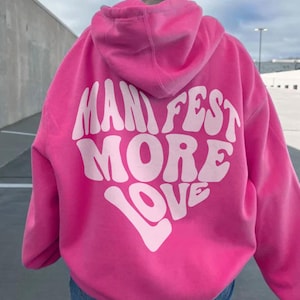 以下が含まれることがあります： レトロなフォントで「MANIFEST MORE LOVE」と書かれた白いハート型のグラフィックがついたピンクのパーカー