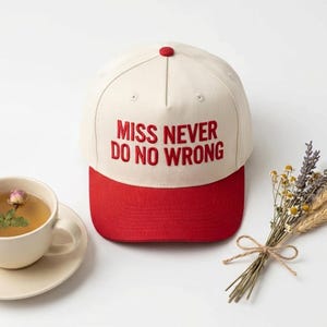 May include: A cream-coloured baseball cap with a red brim and a red button on top. The front of the cap features the embroidered red text "MISS NEVER DO NO WRONG" in a bold, sans-serif font. The cap has a structured five-panel design.
