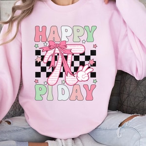 Może przedstawiać: Różowa bluza z nadrukiem "Happy Pi Day" z różową kokardką i szachownicą w tle. Symbol Pi składa się z cyfr Pi.