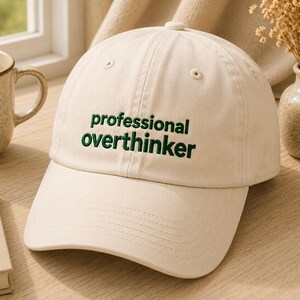 Puede incluir: Una gorra de béisbol de color crema con el texto bordado "professional overthinker" en hilo verde oscuro. La gorra cuenta con una corona estructurada, una visera curva y ojales cosidos en los paneles superiores para ventilación.