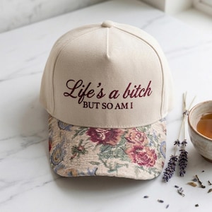 May include: Beige baseball cap with the embroidered text "Life's a bitch BUT SO AM I". The brim features a floral tapestry design with burgundy and pink roses. A stylish and unique accessory.