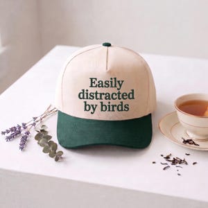 Puede incluir: Una gorra de béisbol con una corona de color crema y una visera de color verde oscuro. La parte delantera de la corona presenta el texto bordado en verde oscuro "Easily distracted by birds" en una fuente serif. Tiene un botón central.