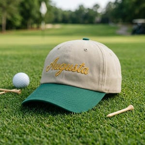 Può includere: Cappellino da baseball beige e verde con la scritta "Augusta" ricamata in oro. Il cappellino è su un campo da golf verde, con una pallina e dei tee nelle vicinanze.