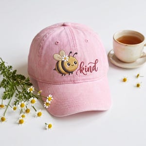 Op de afbeelding: Roze baseballpet met een geborduurde bij en het woord "kind". De bij is geel en bruin met een witte bloem. Een kopje thee en bloemen staan op de achtergrond.