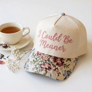 Op de afbeelding: Crèmekleurige baseballcap met de geborduurde zin "I Could Be Meaner" in roze. De klep heeft een bloemen tapestry-ontwerp met blauwe, rode en groene accenten. Een kopje thee en schotel staan op de achtergrond.