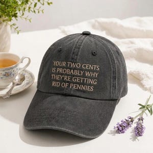 Könnte beinhalten: Eine anthrazitfarbene Baseballkappe mit verwaschenem Finish, die auf der Vorderseite den gestickten Text "YOUR TWO CENTS IS PROBABLY WHY THEY'RE GETTING RID OF PENNIES" in hellem Beige zeigt, mit gebogenem Schirm und einem Knopf oben.