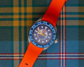 May include: A wristwatch with a bright orange silicone strap and a stainless steel case. The watch face is dark blue with a central logo, white circular hour markers, and yellow hands. A light blue rotating bezel features black numerals.