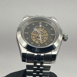 May include: A silver wristwatch with a black face and a gold emblem featuring a crown, anchor, and other symbols. The watch has a silver strap and a textured bezel. The watch is held by a gloved hand.