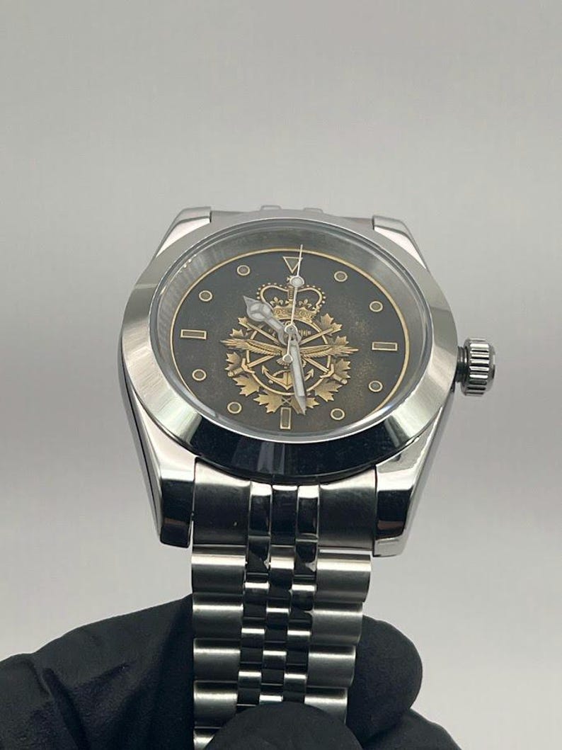 May include: A silver wristwatch with a black face and a gold emblem featuring a crown, anchor, and other symbols. The watch has a silver strap and a textured bezel. The watch is held by a gloved hand.